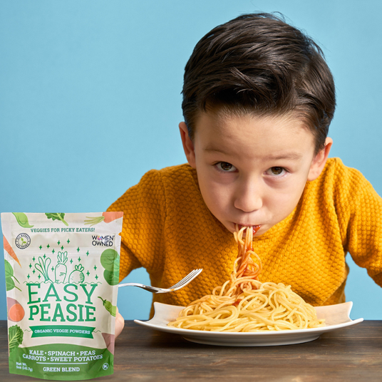 kid with a mouth full of spaghetti , with a pouch of Easy Peasie Green veggie powder next to the plate