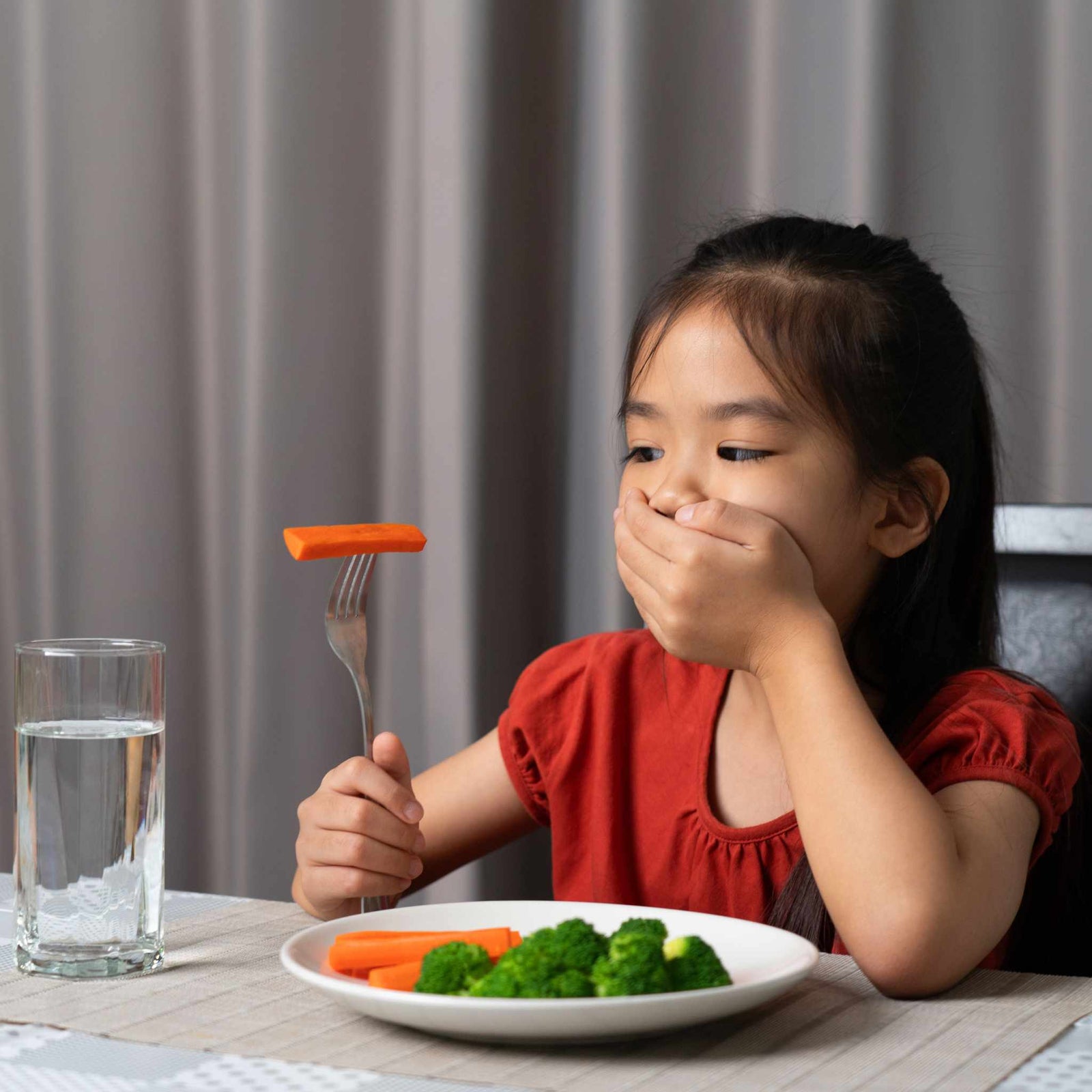 kid covering her mouth and holding a carrot on a fork above a plate of vegetables
